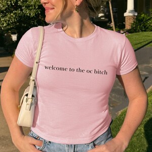 May include: A light pink crew neck t-shirt with the text "welcome to the oc bitch" in black. The shirt is paired with blue jeans. The person is wearing sunglasses and carrying a white purse.