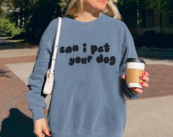 May include: A blue sweatshirt with the text "can i pet your dog" in black. The sweatshirt is worn with black shorts, a white baseball cap, and sunglasses. A white purse hangs from the shoulder.