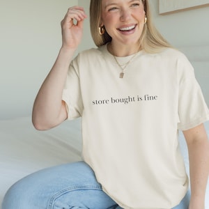 May include: Cream-colored t-shirt with the phrase "store bought is fine" printed in black. The shirt is a relaxed fit and is paired with light blue jeans. The model is smiling and wearing gold jewelry.