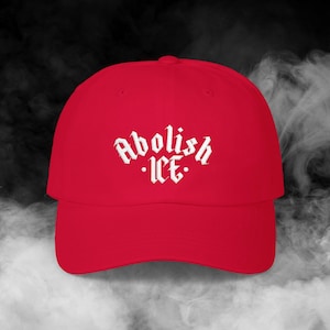 May include: A red baseball cap with the words "Abolish ICE" embroidered in white gothic lettering. The cap has a curved brim and a classic six-panel design. The background is a smoky, dark gray.