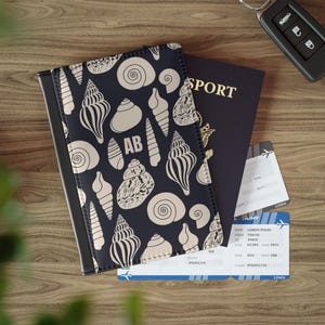 Può includere: Un portapassaporto blu navy con un motivo a conchiglie e le iniziali "AB". Un passaporto, una carta d'imbarco e una chiave dell'auto sono accanto al portapassaporto. Il passaporto ha la scritta "SPORT" in lettere dorate.