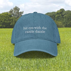May include: A teal baseball cap with the phrase "hit em with the razzle dazzle" embroidered in white. The cap is set against a backdrop of green grass and trees under a blue sky.