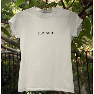 May include: A light beige t-shirt with the words "gay son" printed in black, centered on the front. The shirt is hanging on a clothes hanger against a backdrop of green foliage.