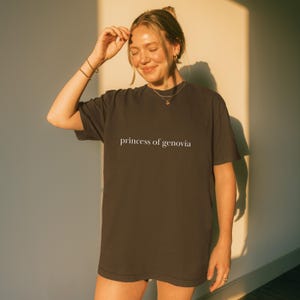 May include: Dark brown t-shirt with the text "princess of genovia" in white. The shirt is a relaxed fit and appears to be made of a soft material. The image is taken in natural light, with a person wearing the shirt.