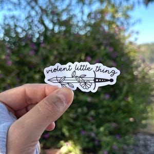 Könnte beinhalten: Ein weißer Aufkleber mit einer schwarzen Strichzeichnung eines Schwertes mit einer Blume auf der Klinge. Der Text "violent little thing" steht über dem Schwert.