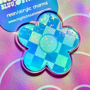 May include: Two flower-shaped resin/acrylic charms. One is a blue flower with a checkered pattern and stars. The other is a blue flower with the text "mystic stickers" and website address. The background is a pastel rainbow.