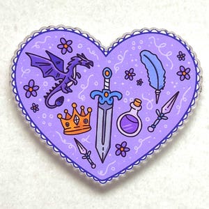 May include: A heart-shaped sticker with a purple background and a scalloped edge. The design features a purple dragon, a sword, a crown, a feather, a potion bottle, and small flowers. The sticker is a fantasy-themed design.