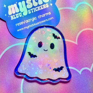 Puede incluir: Un dije de resina holográfica con forma de fantasma con un contorno azul, que presenta una cara sonriente, murciélagos, estrellas y corazones. También hay una tarjeta azul en forma de estrella con el texto "mystic stickers".