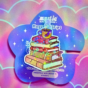 Puede incluir: Una pegatina holográfica con forma de flor que presenta una pila de libros con una taza de café encima. La pegatina tiene el texto "mystic" y "Magsa Grips". La pegatina es compatible con teléfonos y lectores electrónicos.