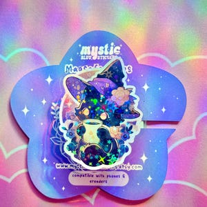 May include: A holographic sticker featuring a cartoon cat wearing a witch hat and holding a mug. The sticker is in a purple flower shape with the text "mystic" and "compatible with phones & ereaders".