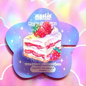 Może przedstawiać: Naklejka przedstawiająca kawałek tortu truskawkowego z bitą śmietaną. Naklejka znajduje się na fioletowym, kwiatowym podkładzie z napisem "mystic blu stickers" i "MagSafe grips". Kompatybilny z telefonami i czytnikami e-booków.