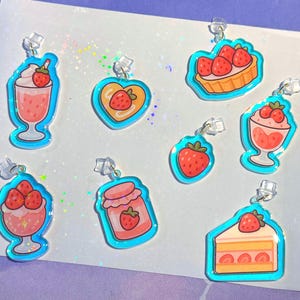 May include: A selection of acrylic charms depicting strawberry-themed treats. The charms include a milkshake, heart design, tart, parfait, single strawberry, jam jar, and cake slice. Each charm has a light blue outline and a silver clasp.