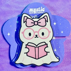 May include: A die-cut sticker featuring a cartoon ghost cat wearing pink glasses and a bow, reading a pink book. The sticker is on a purple star-shaped backing with the text "mystic BLUU STICKERS".