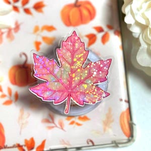 May include: A pink and iridescent maple leaf phone grip. The leaf design features a glittery effect with star-shaped accents. The background includes a fall-themed pattern with pumpkins and leaves.