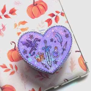 May include: A heart-shaped phone grip with a fantasy theme. The purple grip features a dragon, sword, crown, potion bottle, feather, and daggers. The background is a clear phone case with a fall-themed pattern of pumpkins and leaves.