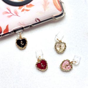 Puede incluir: Cuatro colgantes para teléfono con forma de corazón en negro, blanco, rojo y rosa. Cada colgante tiene un borde dorado y un pequeño diseño de rosa. Los colgantes están unidos a clips de plástico transparente.