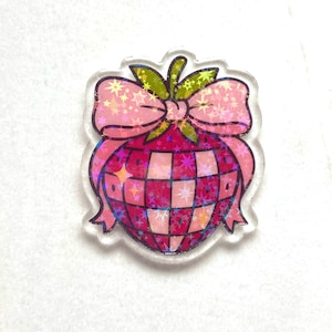 May include: A clear acrylic charm with a pink disco ball design, a bow, and green leaves. The disco ball has a checkered pattern and is surrounded by a pink bow with star accents. A fun accessory.