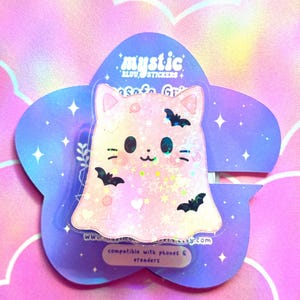 May include: A pink cat-shaped sticker with a ghost-like design, featuring black bat silhouettes and holographic accents. The sticker is on a purple, flower-shaped backing with the text "mystic" and "safe grip".
