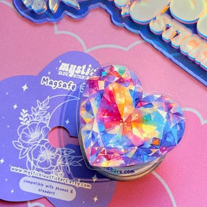 Pu&ograve; includere: Un supporto per telefono MagSafe a forma di cuore con un design a gemma sfaccettata e colorata. L'impugnatura &egrave; su un cartoncino viola con il testo "mysticbluustickers.etsy.com" e "compatibile con telefoni ed e-reader."