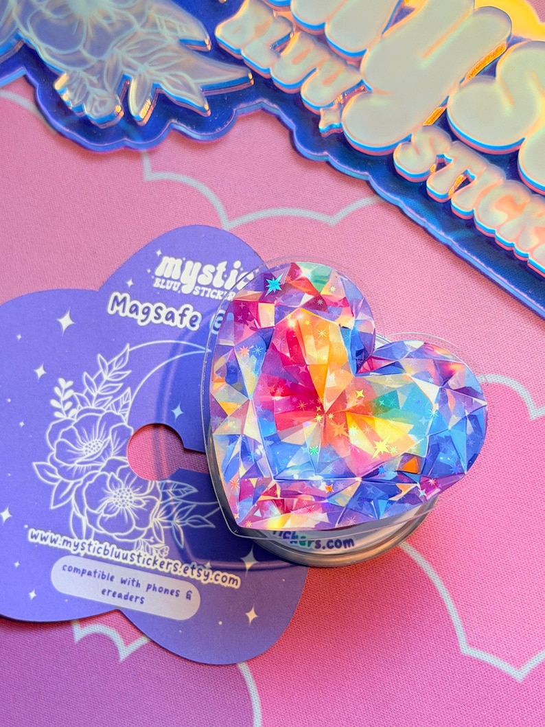 Pu&ograve; includere: Un supporto per telefono MagSafe a forma di cuore con un design a gemma sfaccettata e colorata. L'impugnatura &egrave; su un cartoncino viola con il testo "mysticbluustickers.etsy.com" e "compatibile con telefoni ed e-reader."