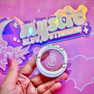 Pu&ograve; includere: Un adesivo circolare trasparente con un bordo bianco &egrave; tenuto davanti a un'insegna olografica che recita "mystic stickers". L'insegna ha un design a forma di luna e floreale. Lo sfondo &egrave; rosa con forme di nuvole.