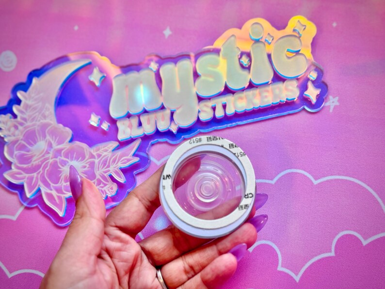 Pu&ograve; includere: Un adesivo circolare trasparente con un bordo bianco &egrave; tenuto davanti a un'insegna olografica che recita "mystic stickers". L'insegna ha un design a forma di luna e floreale. Lo sfondo &egrave; rosa con forme di nuvole.