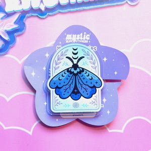 May include: A sticker pack featuring a blue moth design with a crescent moon and star accents, set against a white background within an arched frame. The sticker pack is on a purple, star-speckled, flower-shaped backing, with the text "mystic" at the top.