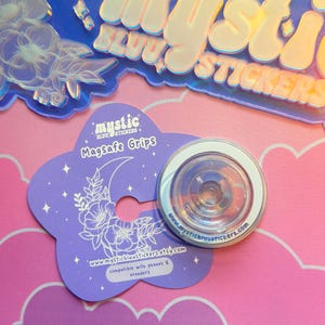Pu&ograve; includere: Un supporto MagSafe trasparente con bordo bianco &egrave; appoggiato su una carta viola a forma di fiore. La carta presenta una luna crescente e un motivo floreale, con il testo "Mystic Stickers" e un indirizzo web. Adesivi olografici sullo sfondo.