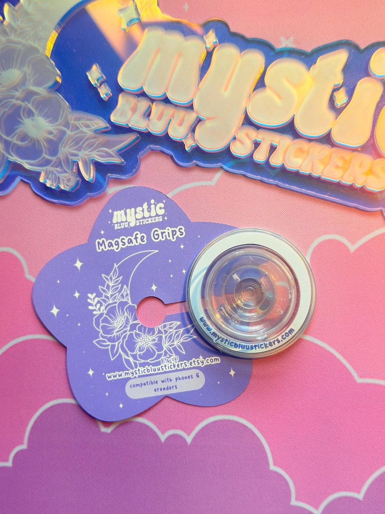 Pu&ograve; includere: Un supporto MagSafe trasparente con bordo bianco &egrave; appoggiato su una carta viola a forma di fiore. La carta presenta una luna crescente e un motivo floreale, con il testo "Mystic Stickers" e un indirizzo web. Adesivi olografici sullo sfondo.