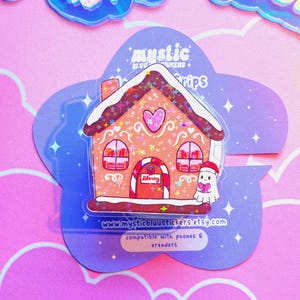 May include: A clear sticker featuring a gingerbread house design with a heart and the word "Library" above the door. The sticker has a ghost wearing a Santa hat and the text "www.mysticbluustickers.etsy.com".