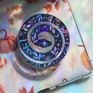 Könnte beinhalten: Ein schwarzer und schillernder Handyhalter mit Spiraldesign. Der Halter hat ein Muster aus Sternen, Monden und Blättern. Der Handyhalter befindet sich auf einer transparenten Handyhülle mit Herbstmotiv.