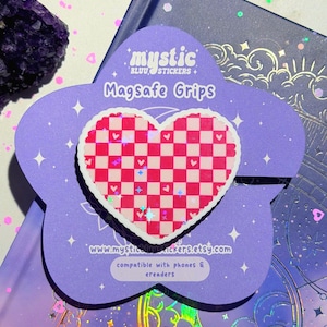 May include: A pink and white checkered heart-shaped MagSafe grip, with a white border, sits on a purple flower-shaped card. The card has the text "mystic BLUU STICKERS + MagSafe Grips" and "compatible with phones & ereaders".
