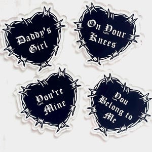 Puede incluir: Cuatro colgantes acrílicos en forma de corazón con texto negro y un borde de alambre de púas. El texto en los colgantes dice "Daddy's Girl", "On Your Knees", "You're Mine" y "You Belong to Me". Los colgantes son transparentes con fondo blanco.