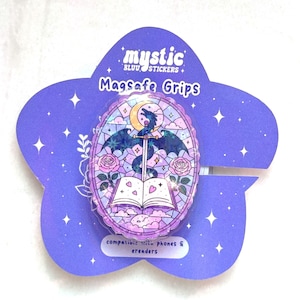 Op de afbeelding: Een paarse Magsafe Grip met een bloemenontwerp en de tekst "mystic". De grip heeft een illustratie in glas-in-loodstijl van een draak, zwaard en open boek. De grip is compatibel met telefoons en e-readers.