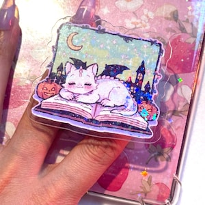 May include: A clear sticker featuring a white cat with bat wings, resting on an open book. The sticker has a Halloween theme with pumpkins, a crescent moon, and a spooky castle in the background. The sticker is held by a person.