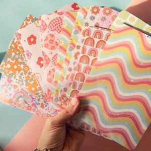 Puede incluir: Un conjunto de 10 hojas de papel de colores con patrones y detalles de purpurina. Los patrones incluyen arco&iacute;ris, corazones, flores y l&iacute;neas onduladas. Las hojas son perfectas para scrapbooking, creaci&oacute;n de tarjetas y otros proyectos de manualidades.