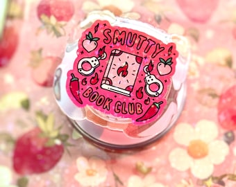 May include: A clear, round sticker with the text "Smutty Book Club" in pink, featuring illustrations of handcuffs, a book with flames, chili peppers, and peaches. The sticker is on a pink floral background with strawberries.