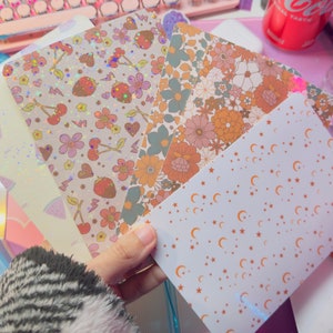 Puede incluir: Cinco hojas de papel decorativo con diferentes patrones. Los patrones incluyen fresas, cerezas, flores y estrellas. El papel es blanco con acentos rosas, naranjas y marrones.