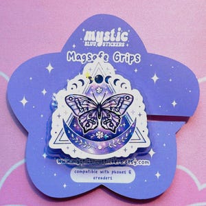 May include: A purple and white butterfly MagSafe grip sticker on a purple flower-shaped backing. The sticker features a butterfly design within a crescent moon and star motif. The text "mystic bluu stickers" and "MagSafe Grips" are visible.