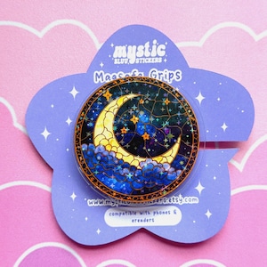 May include: A round phone grip with a stained-glass design of a crescent moon, stars, and clouds. The grip is set against a purple, star-shaped backing with the text "mystic stickers" and "Magsafe Grips". Compatible with phones and e-readers.