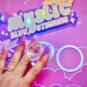 Pu&ograve; includere: Porta adesivi in plastica trasparente con anello bianco e coperchio in plastica trasparente. Lo sfondo presenta un'insegna olografica con la scritta "Mystic Stickers" e uno sfondo rosa.