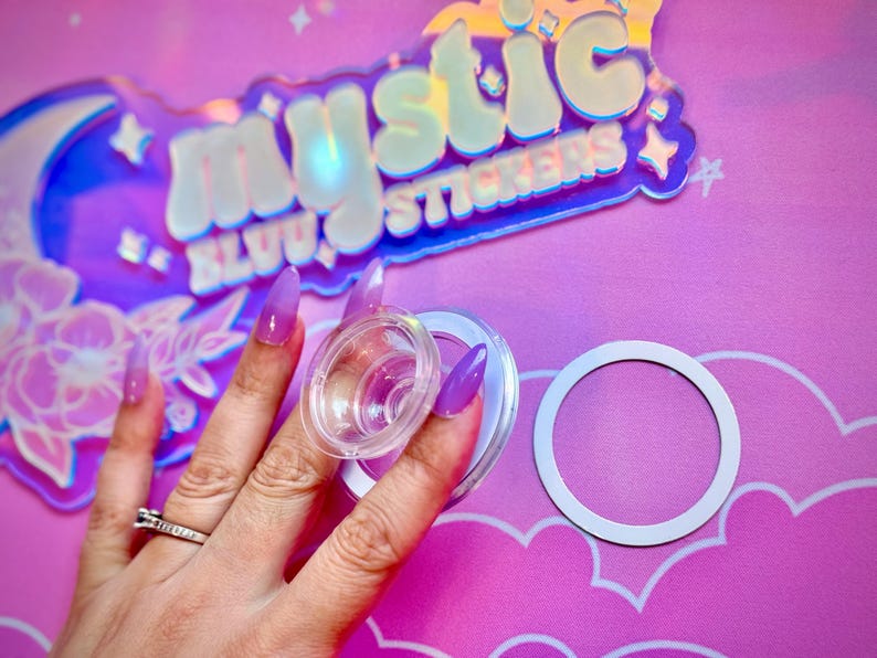 Pu&ograve; includere: Porta adesivi in plastica trasparente con anello bianco e coperchio in plastica trasparente. Lo sfondo presenta un'insegna olografica con la scritta "Mystic Stickers" e uno sfondo rosa.