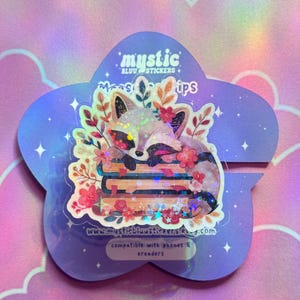 Op de afbeelding: Een holografische sticker met een slapende vos met bloemaccenten, rustend op een stapel boeken. De sticker zit op een paarse, stervormige achtergrond met de tekst "mystic bluu stickers" en "compatible with phones & ereaders".