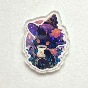 May include: A clear acrylic pin featuring a black cat wearing a purple witch hat adorned with stars and flowers. The cat is holding a mug with a floral design. The pin has a holographic effect and is surrounded by floral and star details.