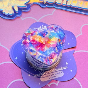 Pu&ograve; includere: Un supporto per telefono trasparente a forma di cuore con un design a gemma sfaccettata e colorata. Il supporto &egrave; posizionato su una base viola a forma di nuvola con il testo "www.mysticbluustickers.etsy.com".