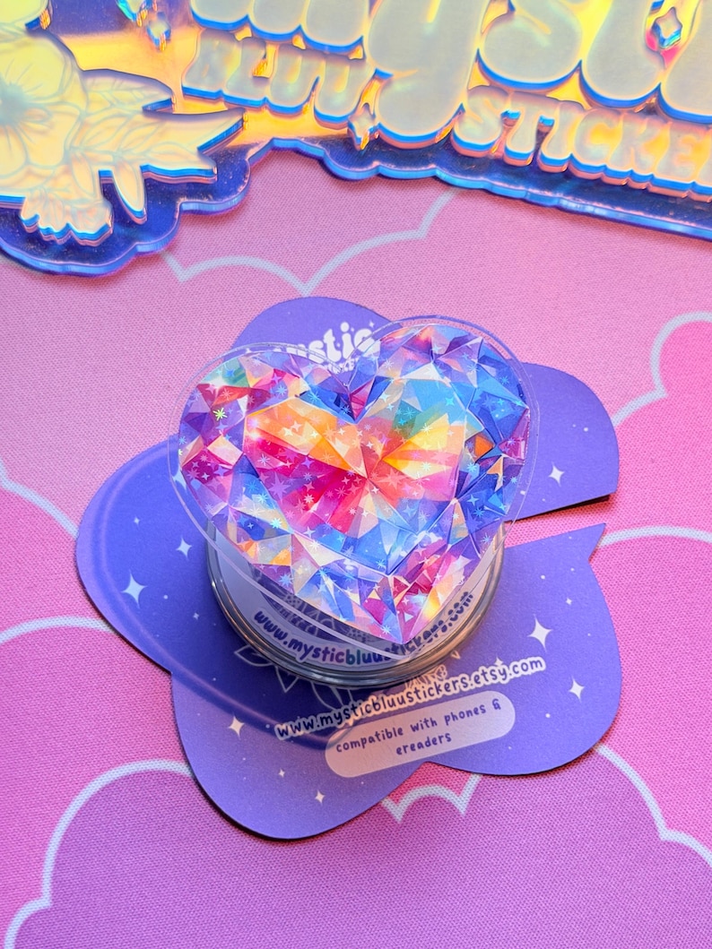 Pu&ograve; includere: Un supporto per telefono trasparente a forma di cuore con un design a gemma sfaccettata e colorata. Il supporto &egrave; posizionato su una base viola a forma di nuvola con il testo "www.mysticbluustickers.etsy.com".