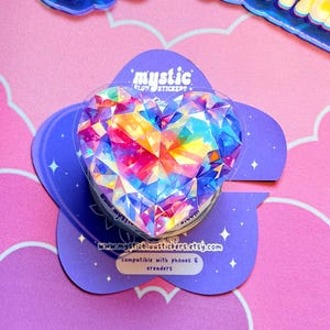 Peut inclure: Un support de téléphone en forme de cœur avec un motif de cristal coloré et facetté. Le support est monté sur une base violette en forme de fleur avec le texte "mystic" et une adresse web. Compatible avec les téléphones et les liseuses.