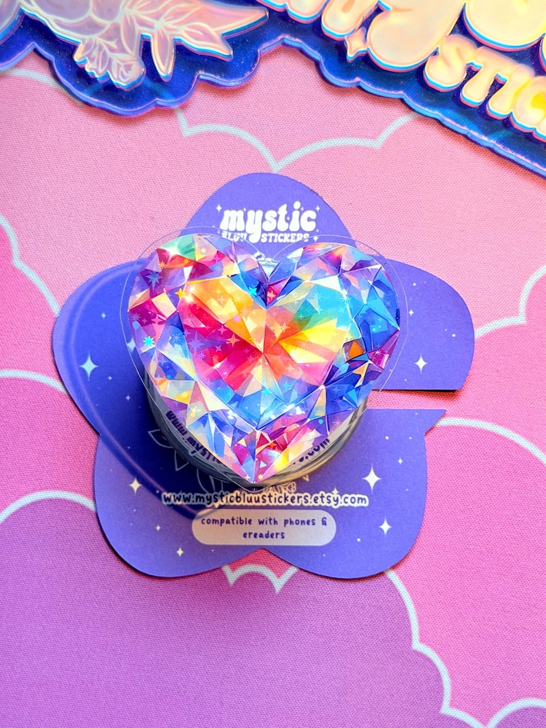 Pu&ograve; includere: Un supporto per telefono a forma di cuore con un design a cristallo sfaccettato e colorato. Il supporto &egrave; montato su una base viola a forma di fiore con il testo "mystic" e un indirizzo web. Compatibile con telefoni ed e-reader.