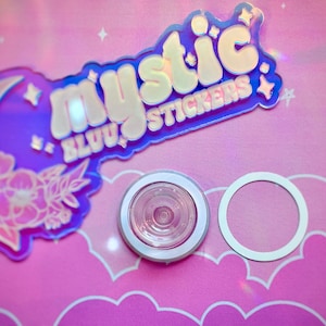 Pu&ograve; includere: Una collezione di adesivi e un piccolo oggetto rotondo con un bordo bianco. Gli adesivi presentano il testo "mystic BLUU stickers" in un design olografico con una luna ed elementi floreali. Lo sfondo &egrave; rosa.