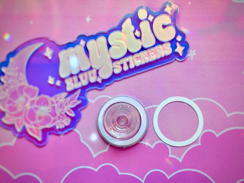 Pu&ograve; includere: Una collezione di adesivi e un piccolo oggetto rotondo con un bordo bianco. Gli adesivi presentano il testo "mystic BLUU stickers" in un design olografico con una luna ed elementi floreali. Lo sfondo &egrave; rosa.
